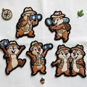 以下が含まれることがあります： 探検家の衣装を着た漫画のシマリスの刺繍パッチ。双眼鏡、カメラ、虫眼鏡を持っています。パッチにはコンパスと葉が含まれています。シマリスは帽子とバックパックを着用しています。