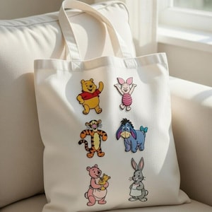 May include: Cream-colored tote bag featuring colorful embroidered images of Winnie the Pooh and friends. The characters include Winnie the Pooh, Piglet, Tigger, Eeyore, Kanga, and Rabbit. The bag has two cream-colored handles.