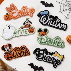 Pode incluir: Seis emblemas bordados com nomes e temas de Halloween. Os emblemas exibem os nomes Emily, Aime, Livia, William, Daniel e Buddy, decorados com ícones que incluem abóboras, um fantasma, Pateta, Minnie Mouse e um osso.
