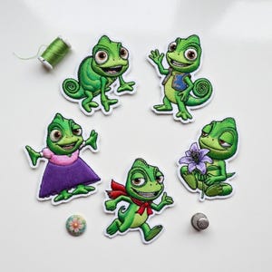 May include: Embroidered patches featuring a green chameleon character in various poses. One patch has a red scarf, another holds a purple flower, and one wears a purple dress. A spool of green thread, a thimble, and a decorative button are also present.