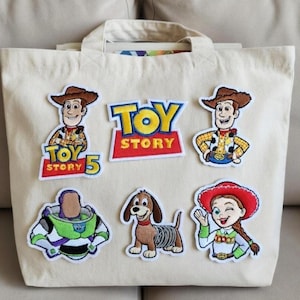 Könnte beinhalten: Eine cremefarbene Tragetasche mit aufgestickten Aufnähern mit Figuren aus dem Animationsfilm "Toy Story". Zu den Aufnähern gehören Woody, Buzz Lightyear, Jessie und Slinky Dog sowie das "Toy Story"-Logo.