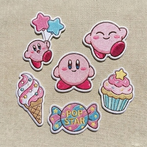 Puede incluir: Una colección de seis parches bordados que incluyen un personaje redondo rosa, un personaje rosa sosteniendo globos en forma de estrella azules y rosas, un helado rosa con estrellas, un cupcake rosa con una estrella y un caramelo con POP STAR.