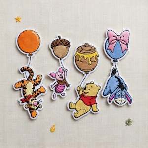 May include: Embroidered patches of Winnie the Pooh characters, including Tigger, Piglet, Winnie the Pooh, and Eeyore. Each character is depicted with a balloon or a honey pot. The patches are brightly coloured with white borders, on a cream-coloured fabric.