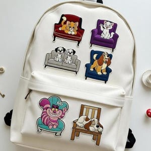 May include: A white backpack adorned with colorful patches featuring cartoon animals on furniture. Patches include a cat on a red chair, a cat on a purple chair, two dogs on a gray couch, a dog on a blue chair, a Cheshire Cat on a teal chair, and a dog on a wooden chair.