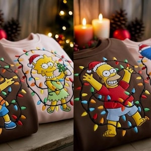 Pode incluir: Close-up de duas camisolas com personagens dos Simpsons. Uma é castanha com Bart, a outra rosa com Lisa. Ambas usam gorros de Pai Natal e estão rodeadas por luzes de Natal. O fundo inclui velas e pinhas.