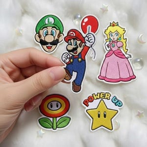 May include: Five embroidered patches featuring characters and icons from the Super Mario franchise: Luigi, Mario holding a red balloon, Princess Peach, a fire flower, and a yellow star with the text Power Up in blue and red lettering.