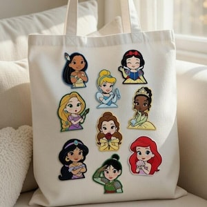 Peut inclure: Un sac fourre-tout en toile blanche orné d'autocollants de princesses Disney. Les autocollants représentent des personnages comme Blanche-Neige, Ariel et Jasmine. Le sac a deux anses et est posé sur une surface blanche.
