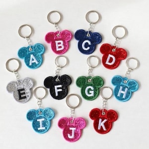 Pode incluir: Uma coleção de chaveiros brilhantes em forma de cabeça de rato, cada um com uma letra do alfabeto. Os chaveiros são em várias cores, incluindo azul, rosa, vermelho, verde e preto, com letras brancas.