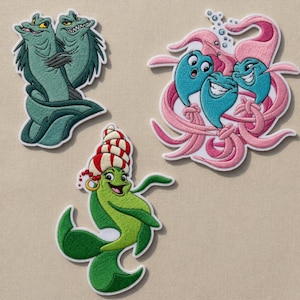 Puede incluir: Tres parches bordados con criaturas marinas de dibujos animados. Un parche muestra dos morenas verdes, otro muestra tres personajes acuáticos azules con tentáculos rosas, y el tercero representa una criatura verde con una concha a rayas rojas y blancas.