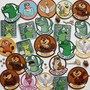 May include: A selection of embroidered patches with cartoon animal designs. The patches feature frogs, beavers, a swan, and a caterpillar. Various shapes are used, including circles and rectangles, with text such as "Tom Lizard" and "Mabel".
