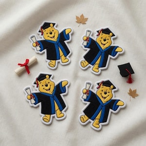 May include: Four embroidered patches of a yellow bear in a graduation gown and cap, holding a scroll. A small mortarboard, a rolled diploma with a red ribbon, and two autumn leaves are also present on a white surface.
