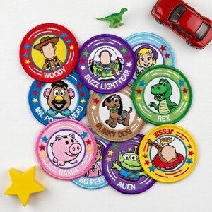 May include: A collection of embroidered patches featuring characters from the animated film "Toy Story." Each circular patch has a different character, including Woody, Buzz Lightyear, and Rex, with their names displayed. A red toy car and a green dinosaur toy are also present.