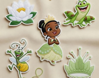Op de afbeelding: Geborduurde patches met een witte waterlelie, een groene kikker, een cartoonprinses in een groene jurk, een groen insect en een groene kroon. De patches liggen op een lichtbeige stof.