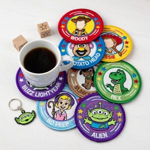 May include: A collection of colorful, round embroidered patches featuring characters from the animated film series, Toy Story. The patches include Woody, Buzz Lightyear, and others. A cup of coffee and a green alien keychain are also present.