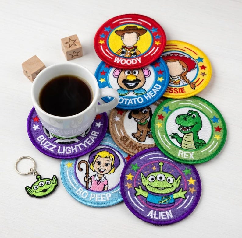 May include: A collection of colorful, round embroidered patches featuring characters from the animated film series, Toy Story. The patches include Woody, Buzz Lightyear, and others. A cup of coffee and a green alien keychain are also present.