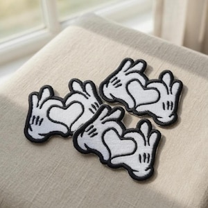 May include: Four white and black embroidered patches in the shape of cartoon hands forming a heart. The patches have a black outline and are arranged on a beige surface. The patches are likely for clothing or accessories.