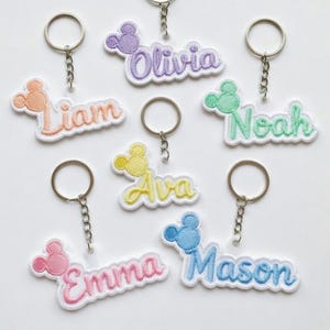 Pode incluir: Chaveiros bordados com nomes como Olivia, Liam, Noah, Ava, Emma e Mason. Cada chaveiro apresenta um design de cabeça do Mickey Mouse em várias cores, com um chaveiro prateado anexado.
