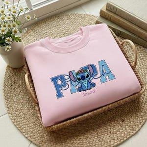 Puede incluir: Sudadera rosa claro con un personaje de dibujos animados azul y la palabra "PAPA" en letras azules. El nombre "Jason" est&aacute; impreso debajo del personaje. La sudadera se muestra en una cesta de mimbre.