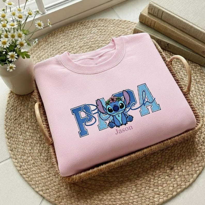 Puede incluir: Sudadera rosa claro con un personaje de dibujos animados azul y la palabra "PAPA" en letras azules. El nombre "Jason" est&aacute; impreso debajo del personaje. La sudadera se muestra en una cesta de mimbre.
