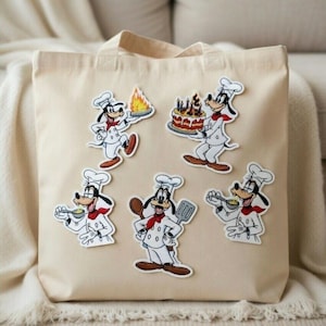 Op de afbeelding: Een beige canvas draagtas versierd met vijf cartoonstickers van een chef-kok. De stickers tonen het personage in een koksmuts en uniform, met diverse kookartikelen zoals een taart, vuur en een lepel. De tas heeft een eenvoudig ontwerp met twee handvatten.