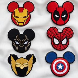 May include: Six embroidered fabric patches shaped like mouse ears, featuring designs inspired by superhero masks and symbols. The collection includes Iron Man, Deadpool, Black Panther, Spider-Man, Thor, and Captain America motifs in red, black, gold, and blue.