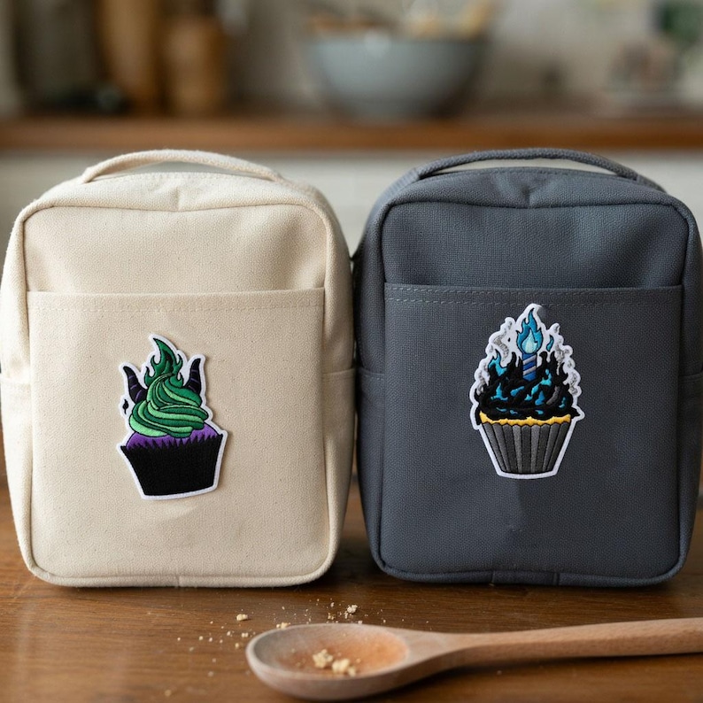 Może przedstawiać: Dwie małe, prostokątne torby. Jedna jest beżowa, druga szara. Każda torba ma przednią kieszeń i naszywkę z babeczką. Naszywka beżowej torby ma zielony lukier i czarne rogi. Naszywka szarej torby ma niebieskie płomienie.