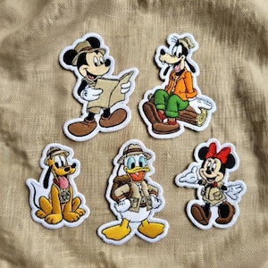 Op de afbeelding: Vijf geborduurde patches met Disney-personages in safari-outfits. Mickey Mouse leest een kaart, Goofy zit op een boomstam, Pluto heeft een rode kraag, Donald Duck draagt een rugzak en Minnie Mouse zwaait. Elke patch heeft een witte rand.