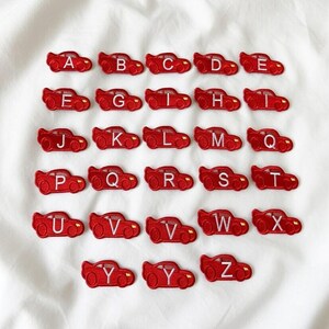 May include: Red car-shaped embroidered patches, each with a different letter of the alphabet from A to Z. The patches are arranged in rows on a white fabric background. The patches are a bright red color.