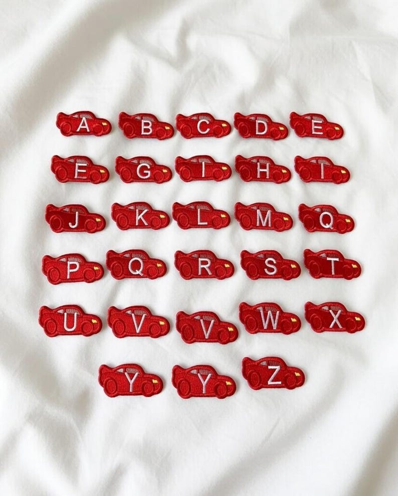 May include: Red car-shaped embroidered patches, each with a different letter of the alphabet from A to Z. The patches are arranged in rows on a white fabric background. The patches are a bright red color.