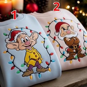 以下が含まれることがあります： 漫画のキャラクターとクリスマスライトが特徴の2つのスウェットシャツ。1つは、サンタ帽と黄色のコートを着たキャラクターが描かれた水色です。もう1つは、サンタ帽と茶色の衣装を着た不機嫌そうなキャラクターが描かれたピンク色です。お祝いのホリデーアパレル。