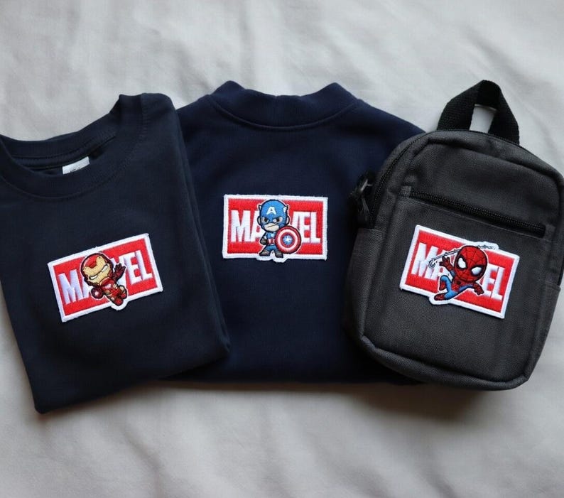 Puede incluir: Tres art&iacute;culos con logotipos de Marvel: una camiseta azul marino con un parche de Iron Man, una sudadera azul marino con un parche del Capit&aacute;n Am&eacute;rica y una bandolera negra con un parche de Spider-Man. Todos los parches est&aacute;n en un rect&aacute;ngulo rojo y blanco.