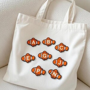 May include: A white tote bag featuring orange and white fish-shaped patches, each with a different letter. The letters are A, B, C, E, G, J, P, and N. The bag has two white handles.
