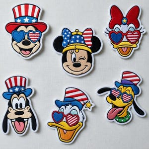 Puede incluir: Parches bordados con Mickey Mouse, Minnie Mouse, Daisy Duck, Donald Duck, Goofy y Pluto. Cada personaje lleva sombreros patrióticos y gafas de sol en forma de corazón con diseños rojos, blancos y azules. Los parches tienen un borde blanco.