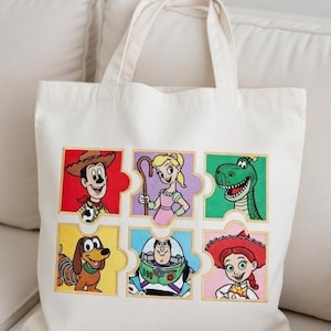 Könnte beinhalten: Beige Canvas-Tragetasche mit bunten, gestickten Bildern von Figuren aus einem beliebten Zeichentrickfilm. Die Bilder sind in einem Puzzle-Muster angeordnet. Die Tasche hat zwei Tragegriffe.
