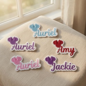 Puede incluir: Cinco parches de tela bordados con nombres y un icono de orejas de ratón. Los parches incluyen los nombres Auriel en azul claro, morado y rosa, Amy en rojo y Jackie en morado. Cada parche tiene un borde blanco y costuras texturizadas.