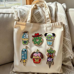 Könnte beinhalten: Cremefarbene Tragetasche mit sechs farbenfrohen Aufnähern im Cartoon-Stil. Zu den Aufnähern gehören ein Eisbecher, ein Donut, eine Figur mit Mäuseohren, ein Eis am Stiel, ein Cupcake und eine Figur in einer Eistüte.