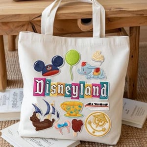 May include: Off-white canvas tote bag with embroidered Disneyland patches. Patches include Mickey Mouse ears, a green balloon, Dumbo the elephant, a cupcake, a ship, a teacup, a train, a bow, a Mickey Mouse ice cream bar, and a "it's a small world" medallion.