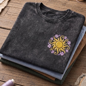Könnte beinhalten: Ein schwarzes T-Shirt in Acid-Wash-Optik mit einem gestickten kreisförmigen Design auf der Brust. Die Stickerei zeigt eine goldgelbe Sonne mit wirbelnden Strahlen, umgeben von kleinen violetten Blumen. Das T-Shirt hat einen Rundhalsausschnitt.