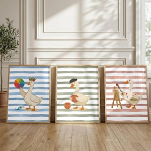 May include: Three framed wall art prints featuring white geese on striped backgrounds. The first goose holds a color wheel, the second wears a beret and holds a pot, and the third stands by an easel with a paintbrush and a paint palette.