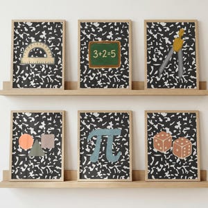 May include: Six framed rectangular prints displayed on two wooden shelves. Each print features a black and white speckled background. The illustrations include a protractor, a chalkboard with 3+2=5, a compass, geometric shapes, a pi symbol, and two dice.