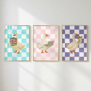 May include: Three framed wall art prints featuring white geese on checkered backgrounds. The first goose holds a stack of books on a turquoise grid, the second uses a laptop on a pink grid, and the third holds colorful geometric shapes on a purple grid.