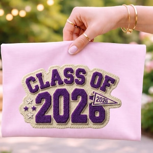 Pode incluir: Um item de tecido rosa claro com o texto "CLASS OF 2026" em roxo, delineado em bege claro. Duas pequenas estrelas e uma flâmula também estão incluídas no design. O item está sendo segurado por uma pessoa.