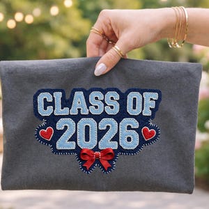 Op de afbeelding: Een grijze clutch met de tekst "CLASS OF 2026" in blauw en wit, met rode harten en een rode strik. De clutch wordt omhoog gehouden door een hand met gouden ringen en armbanden, met een vage achtergrond van groen en lichten.