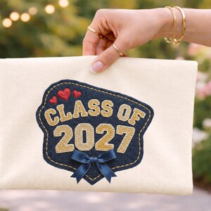Op de afbeelding: Crèmekleurige clutch met een marineblauwe en gouden geborduurde patch. De patch leest "CLASS OF 2027" met een blauwe strik en twee rode hartjes. De clutch wordt vastgehouden door een persoon met gouden sieraden.