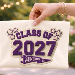 Pode incluir: Tecido creme com um design bordado roxo que diz "CLASS OF 2027 SENIORS". O design inclui estrelas e uma faixa com o ano de 2027. O tecido é segurado por uma mão com joias de ouro.