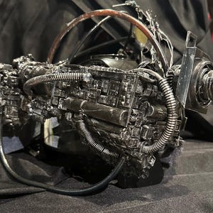 May include: A detailed, metallic, futuristic device with a complex arrangement of tubes, wires, and mechanical components. The device has a prominent lens with a red center and a curved, arched structure. The overall color scheme is dark gray and black.
