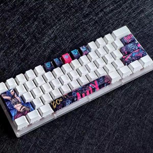 Op de afbeelding: Een witte mechanische toetsenbord met aangepaste keycaps. De keycaps hebben kleurrijke ontwerpen, waaronder een karakterillustratie en graffiti-kunst in roze, blauw en zwart. Sommige toetsen hebben tekst en symbolen. Het toetsenbord ligt op een donkere ondergrond.