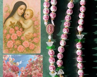 May include: A rosary featuring white beads with pink floral patterns, gold-tone spacers, and clear glass flower accents. It includes a pink oval centerpiece with a religious figure and a gold-tone cross. Text reads: St Theresa the Little Flower.