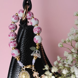 Könnte beinhalten: Ein dekorativer Perlenanhänger mit weißen Keramikperlen mit rosa Blumenmuster, unterbrochen von kleinen goldfarbenen Zwischenstücken. Der Anhänger enthält eine transparente glockenförmige Perle, einen ovalen religiösen Anhänger und ein goldenes Kreuz mit Perlen.