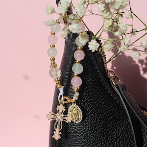 Könnte beinhalten: Ein dekorativer Taschenanhänger mit einer Reihe aus rosa und blassgrünen Perlen, die durch kleine goldfarbene Zwischenelemente getrennt sind. Der Anhänger enthält ein goldfarbenes Kreuz mit winzigen weißen Perlen und eine kleine ovale Medaille.